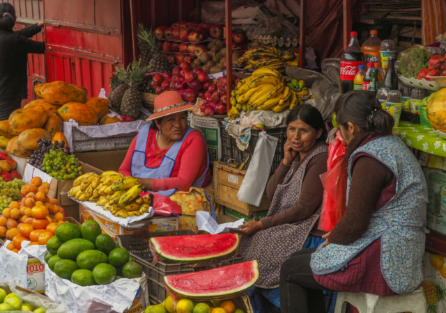 Three women chat at a fruit stall, surrounded by bananas, watermelons, papayas, and other produce.