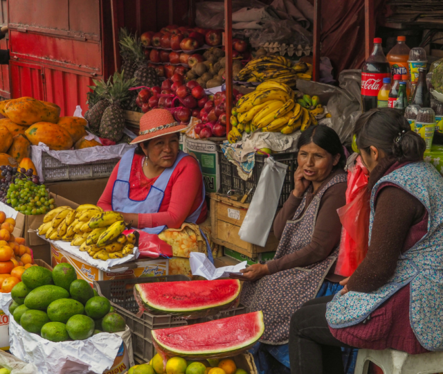 Three women chat at a fruit stall, surrounded by bananas, watermelons, papayas, and other produce.